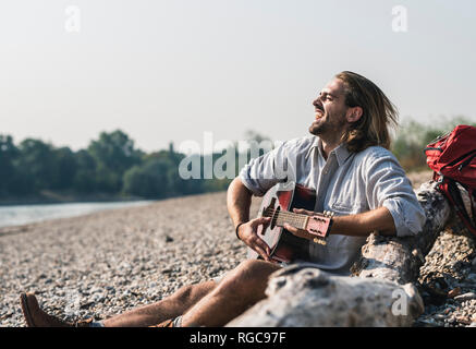 Ausgelassene junge Mann am Flußufer Gitarre spielen Sitzen Stockfoto