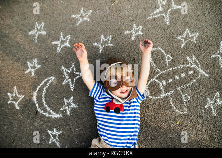Portrait von lächelnd Kleinkind tragen pilot Hut und Brille liegen auf Asphalt mit Flugzeug, Mond und Sternen bemalt Stockfoto