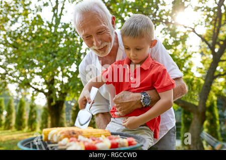 Großvater, Enkel drehen einen Maiskolben beim Grillen im Garten Stockfoto