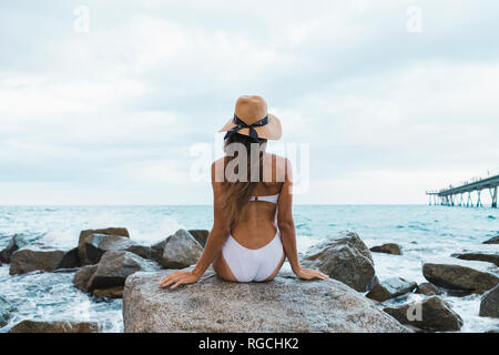 Ansicht der Rückseite des jungen Frau das Tragen von Badebekleidung und Hut sitzt auf Felsen im Meer Stockfoto