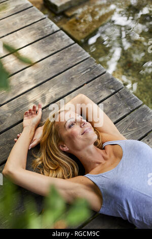 Lächelnde blonde Frau liegend auf hölzernen Steg am See Stockfoto