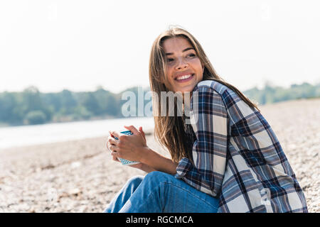 Glückliche junge Frau mit einem Becher am Fluss sitzen Stockfoto