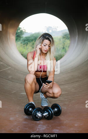 Athletische Frau zusammengekauert in einer Röhre mit Hanteln und Kopfhörer an Zelle Telefon Stockfoto