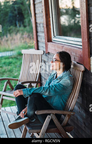Glückliche junge Frau sitzt auf der Veranda eines Holzhauses Stockfoto