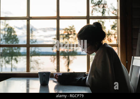 Finnland, Lappland, junge Frau am Fenster an einem See in Tagebuch suchen Sitzen Stockfoto