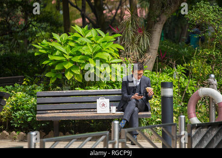 Ein Mann im Anzug sitzt auf einer Bank im Hong Kong Park, hält ein Sandwich an der Hand für sein Mittagessen im Freien, um seinen Geist zu tanken und zu entspannen. Hongkong. Stockfoto