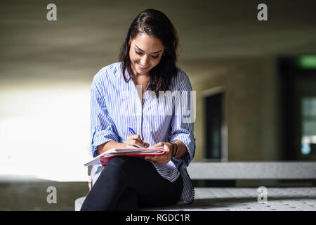 Smiling Student sitzt auf der Bank im Freien Notizen in einem Notebook Stockfoto