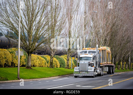 Klassische Motorhaube kommerziellen amerikanischen Big Rig Semi Truck mit Flachbett Auflieger für den Transport von Containern mit Glas fahren auf der Straße mit Bäumen und Stockfoto