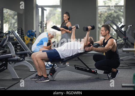 Passen die Leute trainieren in Modrn Fitnessraum. Starke Mann lag auf der Werkbank, Hände hoch halten mit Hanteln. Personal Trainer ihn unterstützen. Weiblichen Trainer helfen, alter Mann mit heraus zu arbeiten. Stockfoto