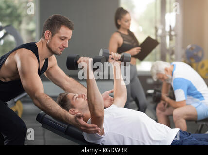 Seitenansicht der Mann im weißen T-Shirt liegen auf der Werkbank, holding hands up mit Hanteln. Muskulöse Trainer helfen Client mit sportlicher Ausstattung. Konzept der modernen Fitnessraum und Sport. Stockfoto