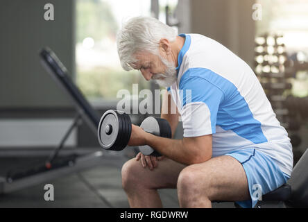 Gesunde ältere Menschen Übungen im modernen Fitnessraum, über sportliche Ausstattung. Fit und sportlich Mann sitzt auf besondere Sitzbank, lehnte sich auf Knie. Rentner, hartnäckig zu arbeiten. Stockfoto