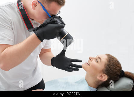 Wunderschöne Frau liegend auf Zahnarztstuhl und posieren. Adorable, schöne Frau, Zähne und strahlendes Lächeln nach Zahnmedizin Pflege. Fotograf in schwarz Handschuhe mit Foto. Stockfoto