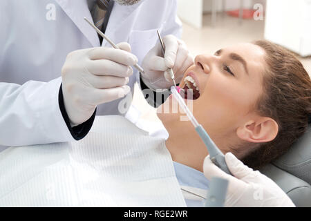 Schöne Frau im Zahnarztstuhl liegen und in ihre Zähne getan. Ärzte in weißen Handschuhen holding Restaurierung Instrumente im Hohlraum und Härtung der Zähne. Konzept der Zahnheilkunde. Stockfoto