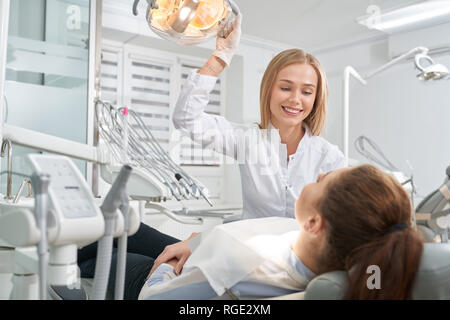 Patient liegt auf Zahnarztstuhl und Rücksprache mit dem Arzt. Schöne Frau in weißer Uniform und Handschuhe Hand auf speziellen Lampe, lächelnd, am Client. Stockfoto