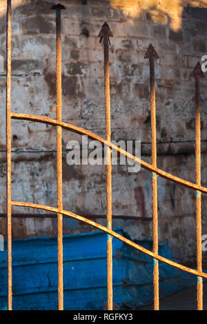 Verrostete Zaun in Form von Pfeilen, erleuchtete durch die Morgensonne. Steinmauer und einem blauen defektes Stück eines anderen Zaun im Hintergrund. Hafen von Essaouir Stockfoto