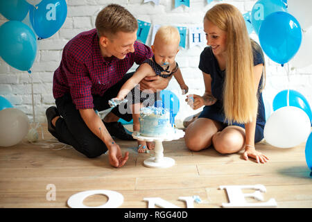 Eltern sind feiert ersten Geburtstag Ihres einjährigen Sohn und Fütterung seiner Durch festliche Kuchen auf dem Hintergrund der blauen Luftballons. Stockfoto