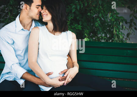 Eine junge moderne Paar sitzt auf einer Bank mit Blätter im Hintergrund, dass sie schwanger ist und Wiegen ihre runden Bauch sanft. Er umarmt sie zärtlich. Stockfoto