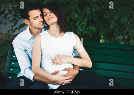 Eine junge moderne Paar sitzt auf einer Bank mit Blätter im Hintergrund, dass sie schwanger ist und Wiegen ihre runden Bauch sanft. Er umarmt sie zärtlich. Stockfoto