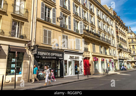 Ein Mann und eine Frau mit einem kleinen Kind auf einem Roller vorbei gehen die Geschäfte und Wohnhäuser auf der Rue Saint-Dominique, Paris, Frankreich Stockfoto