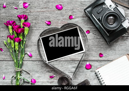 Retro Kamera und leeren Bilderrahmen in Herzform Film mit Purple carnation Blüten, Blütenblätter und gefütterte Notebook mit rustikalen Holzmöbeln Hintergrund. Valentin Stockfoto