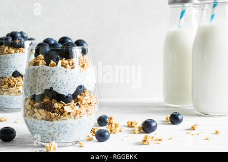 Chia Pudding oder Joghurt Parfait mit Blaubeeren, Müsli und Chia Samen auf weißem Hintergrund aus Holz mit Flaschen Milch. gesundes Frühstück Stockfoto