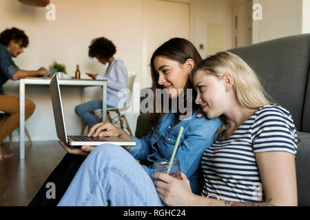 Zwei lächelnde junge Frauen sitzen auf dem Boden teilen Laptop mit Freunden im Hintergrund Stockfoto