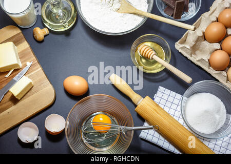 Küchengeräte und Zutaten für Kuchen backen. Eier, Mehl, Öl, Butter, Milch, Honig, Zucker und Schokolade auf dunklem Hintergrund. Backen Konzept, Anzeigen Stockfoto