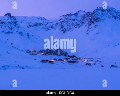 Schnee bedeckt Tignes Val Claret vom See in der Abenddämmerung mit rosa Himmel hinter Blue Mountain Range mit Straßenbeleuchtung und eine Person zu Fuß Hund über See Stockfoto