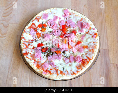 Vorbereitet appetitliche Pizza mit Speck, Käse und Gemüse auf runde Platte auf Sandy braun Küche aus Holz Tisch Ansicht Nahaufnahme Stockfoto