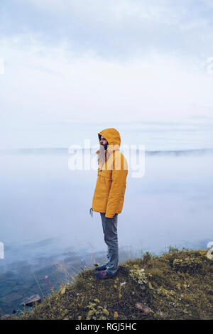 Schweden, Lappland, Mann mit Vollbart tragen gelbe Windbreaker stehend an Water's Edge auf Distanz suchen Stockfoto