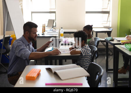 Seitenansicht der jungen Lehrer helfen jungen mit Studie während kniend vor seinem Schreibtisch im Klassenzimmer Stockfoto