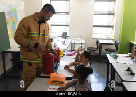 Seitenansicht der männlichen Kaukasier Feuerwehrmann Lehre schoolkids über Brandschutz, Feuerlöscher in Klassenzimmer der Volksschule Stockfoto