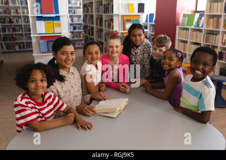 Vorderansicht des gerne Lehrerin und schoolkids an Kamera suchen während des Studiums am Tisch in der Bibliothek Stockfoto