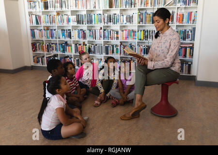 Seitenansicht der weiblichen Lehrer sitzen auf einem Stuhl und lesen eine Geschichte schoolkids sitzen auf dem Boden in der Schule Bibliothek Stockfoto