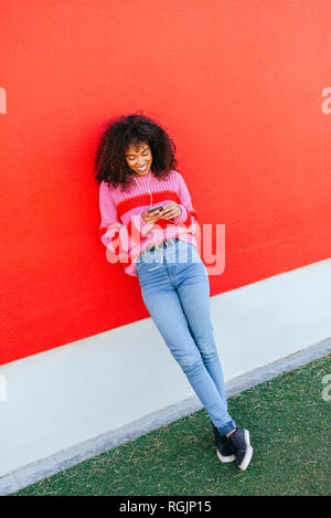Lächelnde junge Frau mit Kopfhörer gegen rote Wand an der Zelle Telefon schiefen Stockfoto