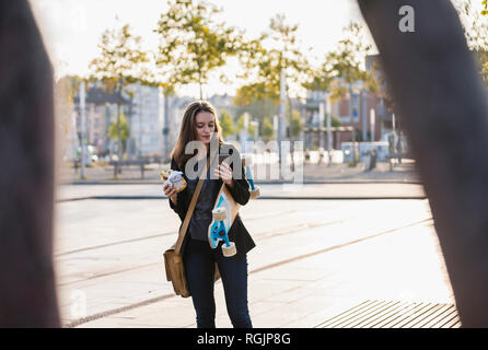 Junge Frau mit Longboard und Snack in der Stadt prüfen Handy Stockfoto