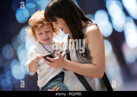 Mutter trägt ihren kleinen Sohn und sie lächeln, als sie ihm zeigt ein Handy. Stockfoto