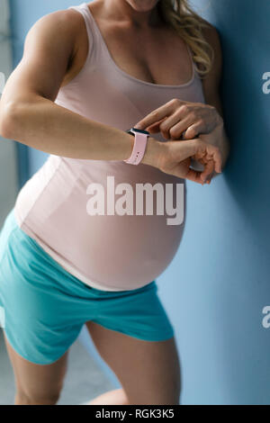 Im mittleren Abschnitt der schwangeren Frau, die auf der blauen Wand betrachten Smartwatch
