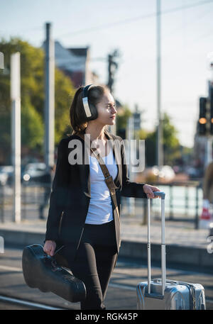 Junge Frau mit Kopfhörer, Koffer und Violine bei tram station Stockfoto