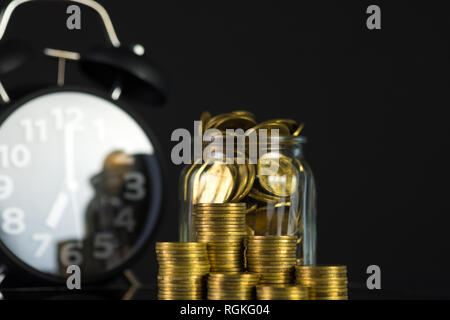 Münzen Stapeln mit Münze in Glas Glas Flasche und Wecker in dunklen Zimmer, Business und Finanzen Konzept Idee. Stockfoto