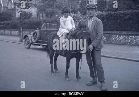 1920s, historische Bild, auf dem ein junges Mädchen auf einem Pony oder Pferd in einer kleinen Straße, geführt von einem stalljungen, England, UK. Ein Kraftfahrzeug der Epoche gesehen werden kann auf der Seite der Straße geparkt. Stockfoto