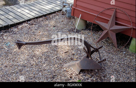 Antike eiserne Pflug sitzen auf Kieselsteinen in der Rückseite eines roten Holzhaus mit einem Bügeleisen Texas Stern lehnte sich auf dem Haus. Stockfoto