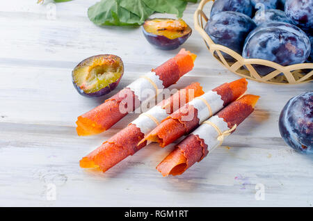 Frisches Lila Pflaumen in einem korbwaren Vase und auf dem Tisch verstreut, Pflaumen in Form von trockenen Späne und Obst Leder rollt auf weißen Holz- Hintergrund. D Stockfoto