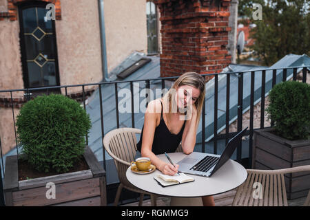 Lächelnde junge Frau sitzt auf dem Balkon mit dem Laptop von Notizen Stockfoto
