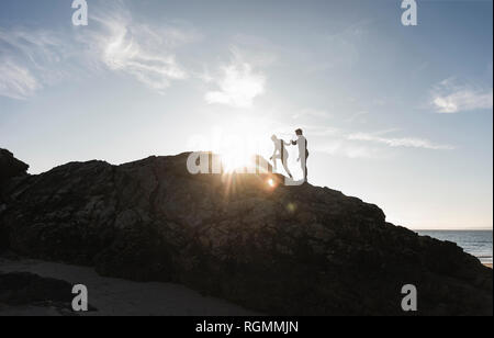 Frankreich, Bretagne, junges Paar klettern auf einem Felsen am Strand bei Sonnenuntergang Stockfoto