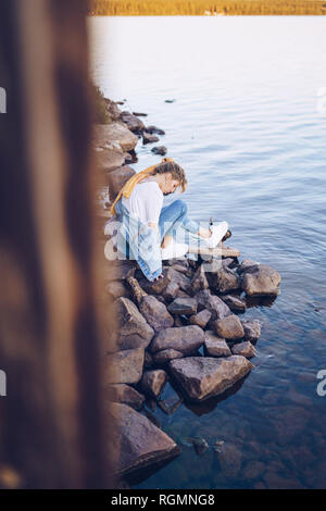 Schweden, Lappland, nachdenkliche junge Frau im Water's Edge sitzen Stockfoto