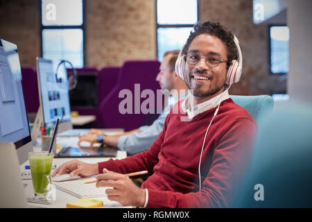 Junger Mann am Schreibtisch im Büro sitzen, tragen, Kopfhörer, lächeln Stockfoto