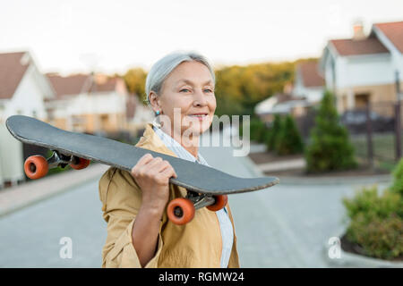 Portrait von lächelnden älteren Frau mit Skateboard auf ihrer Schulter Stockfoto