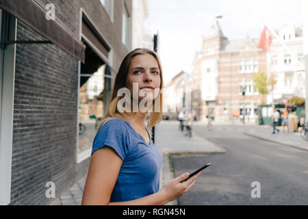 Niederlande, Maastricht, lächelnde junge Frau mit Handy in der Stadt umher, um die