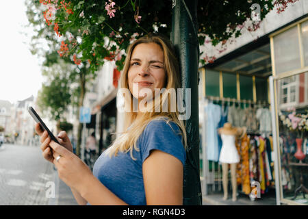 Niederlande, Maastricht, lächelnde junge Frau mit Handy in der Stadt funkeln